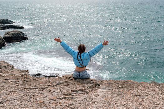 A woman in a blue jacket sits on a rock above a cliff above the sea, looking at the stormy ocean. Girl traveler rests, thinks, dreams, enjoys nature. Peace and calm landscape, windy weather