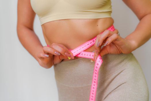 Cropped view of slim woman measuring waist with tape measure at home, close up. An unrecognizable European woman checks the result of a weight loss diet or liposuction indoors. Healthy lifestyle