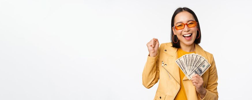 Microcredit and money concept. Stylish asian young woman in sunglasses, laughing happy, holding dollars cash, standing over white background.