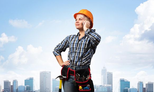 Attractive blonde woman in workwear and hardhat talking on smartphone. Portrait of young worker in checkered blue shirt standing on cityscape background. Business communication and conversation.