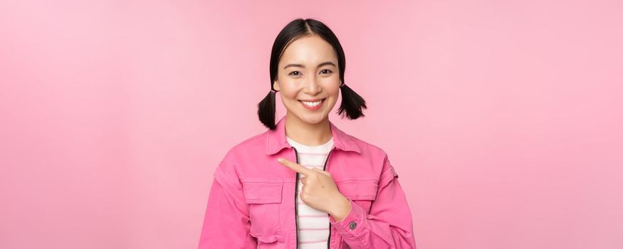 Portrait of beautiful smiling asian girl, pointing finger left, showing advertisement, banner promo, standing over pink background.