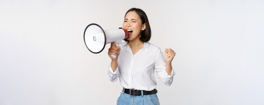 Enthusiastic asian woman, girl activist shouting at protest, using megaphone, looking confident, talking in loudspeaker, protesting, standing over white background.