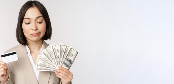 Thoughtful businesswoman, korean corporate woman showing credit card and money cash, dollars in hands, standing over white background and thinking.