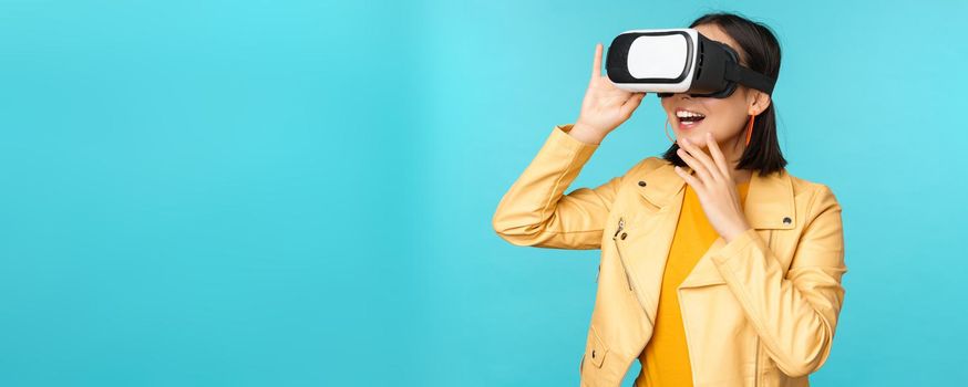 Portrait of happy asian female model using VR headset, smiling and laughing in virtual reality glasses, standing over blue background.