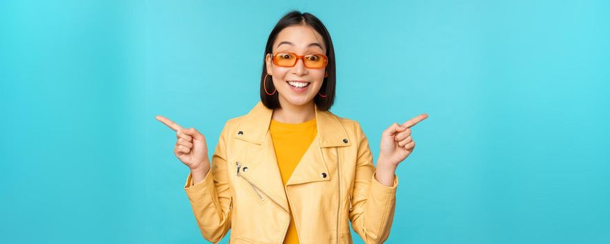Happy stylish chinese girl in sunglasses, points fingers sideways, invites to check out, see store discount, shows left and right, stands over blue background.