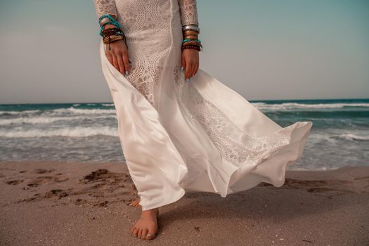 Model in boho style in a white long dress and silver jewelry on the beach. Her hair is braided, and there are many bracelets on her arms