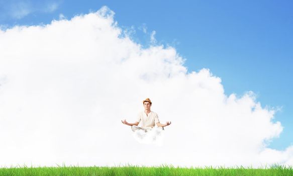 Young man keeping eyes closed and looking concentrated while meditating on cloud in the air with bright and beautiful landscape on background.