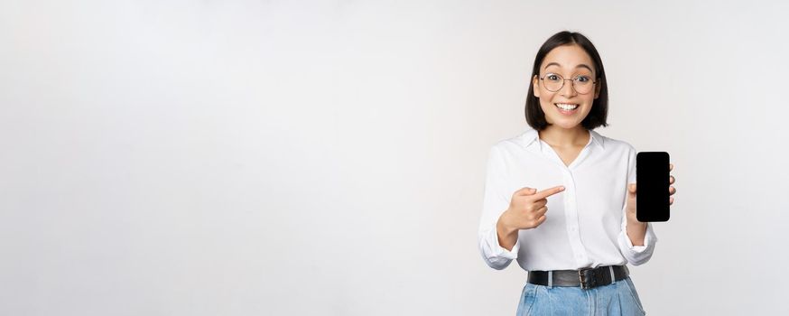 Enthusiastic young asian woman pointing finger at smartphone screen, showing advertisement on mobile phone, white background.