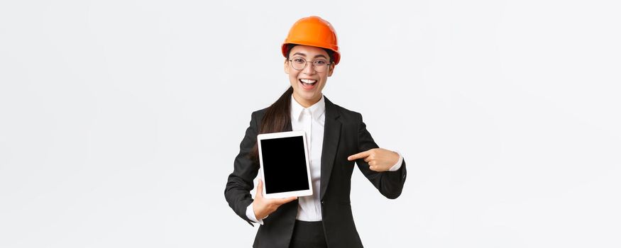 Smiling professional female construction manager, asian engineer in safety helmet and business suit show project, pointing finger at digital tablet screen with pleased expression, white background.