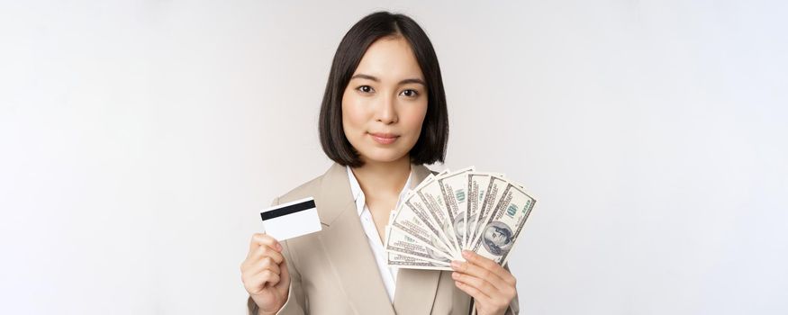 Close up of asian businesswoman, office lady showing credit card and money dollars, standing in suit over white background.