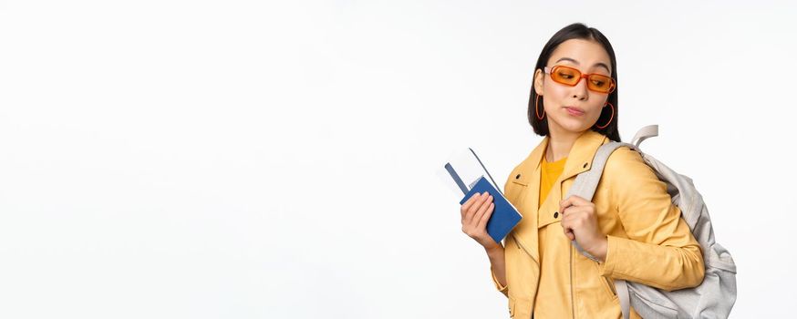 Stylish asian woman in sunglasses going abroad with backpack, tourist holding passport and flight boarding tickets, standing over white background. Copy space