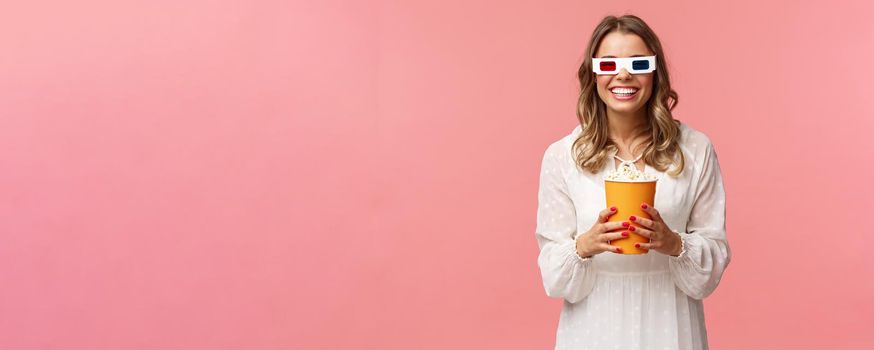 Leisure, going-out and spring concept. Portrait of happy carefree blond girl in white dress enjoying watching movie in 3d, wearing glasses eating popcorn and smiling, attend cinema, pink background.