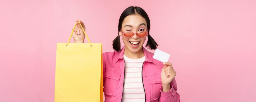 Happy young asian woman showing credit card for shopping, holding bag, buying on sale, going to the shop, store, standing over pink background.