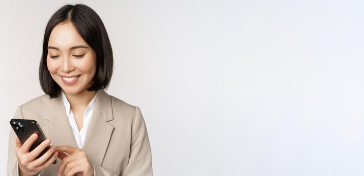 Close up portrait of korean woman, corporate lady in suit, using mobile phone and smiling, holding smartphone, standing over white background.