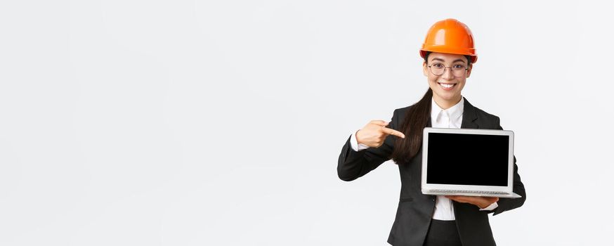 Smiling pleased asian female chief engineer showing chart with enterprise profit, making presentation during investors meeting, wearing safety helmet, pointing at laptop screen, white background.