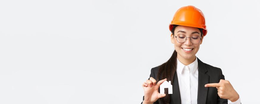 Close-up of smiling asian female engineer, architect in helmet and business suit holding home miniature and pointing finger at small house, working over building construction, white background.