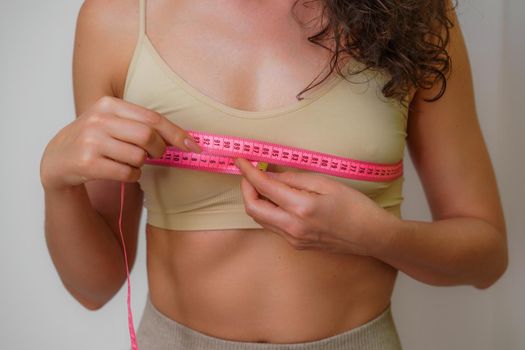 Cropped view of slim woman measuring breasts with tape measure at home, close up. Unrecognizable European woman checks the result of a weight loss diet or liposuction indoors. Healthy lifestyle..