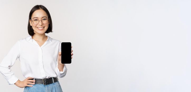 Smiling korean woman showing smartphone screen, demonstrating mobile application, standing over white background.