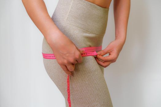 Cropped view of slim woman measuring hips with tape measure at home, close up. An unrecognizable European woman checks the result of a weight loss diet or liposuction indoors. Healthy lifestyle