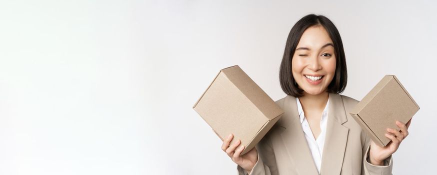 Image of saleswoman, asian businesswoman holding boxes with company brand product, smiling at camera, standing against white background.