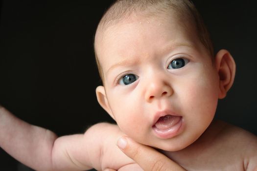 Newborn baby against black background with big eyes looking into the camera being held by her mother