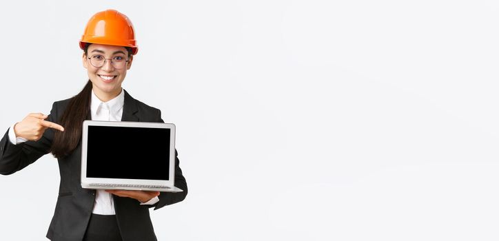 Smiling pleased asian female chief engineer showing chart with enterprise profit, making presentation during investors meeting, wearing safety helmet, pointing at laptop screen, white background.