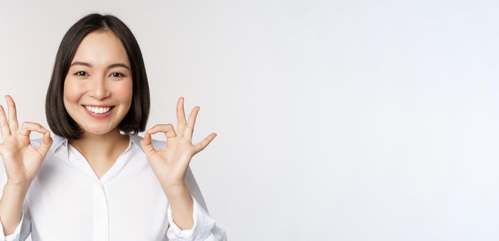 Close up head portrait of asian girl showing okay, ok sign and smiling satisfied, recommending, being pleased, praise and make compliment, white background.