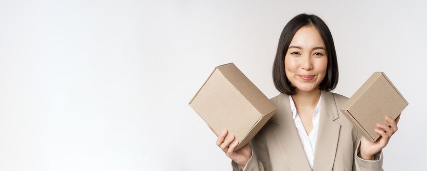 Image of saleswoman, asian businesswoman holding boxes with company brand product, smiling at camera, standing against white background.