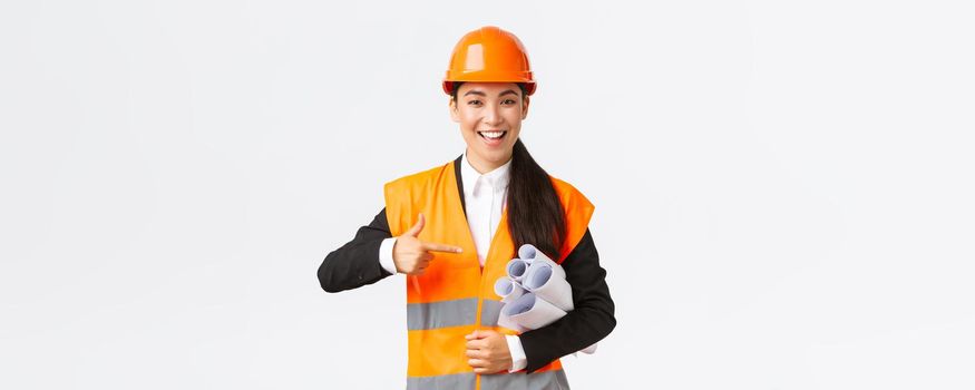 Professional confident asian female architect, chief engineer in safety helmet pointing finger at blueprints, showing project plan or documents for construction, standing white background.