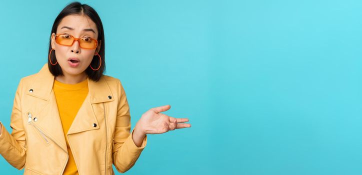 Portrait of chinese girl in sunglasses, looks confused, shrugs shoulders and staring puzzled at camera, standing over blue background.