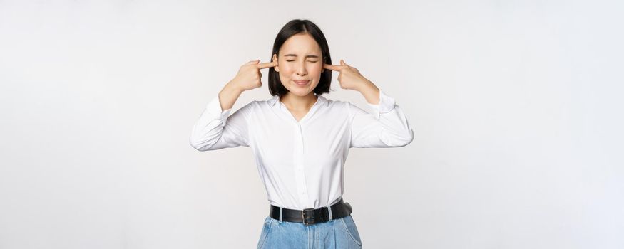 Annoyed asian woman feeling discomfort from loud annoying noise, close ears and eyes, standing over white background. Copy space