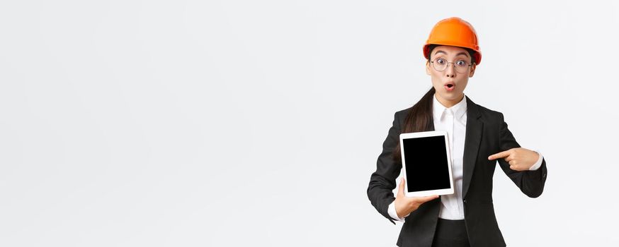 Impressed and surprised asian female engineer, technician at industrial area wear safety helmet and business suit, pointing finger at profit chart, diagram at digital tablet screen, look amazed.