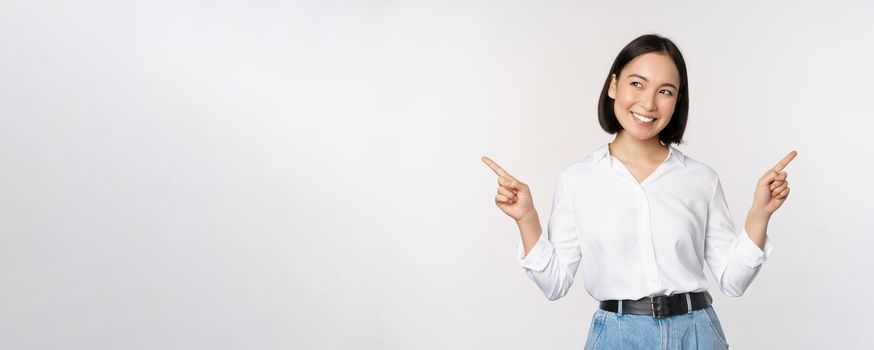 Image of beautiful asian woman pointing fingers left and right, making decision, showing two variants choices, standing over white background.