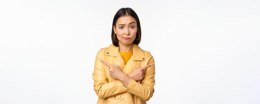 Image of indecisive asian girl, pointing fingers sideways, pointing left and right, choosing variant, deciding, standing over white background. Copy space