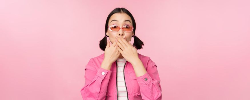 Portrait of shocked, stylish asian girl in sunglasses, closes mouth, looks with surprised face expression, stands over pink background. Copy space