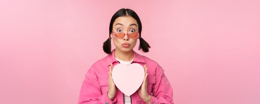 Cute asian girl showing heart shaped box with gift, looking surprised and excited, romantic present concept, wearing sunglasses, standing over pink background.