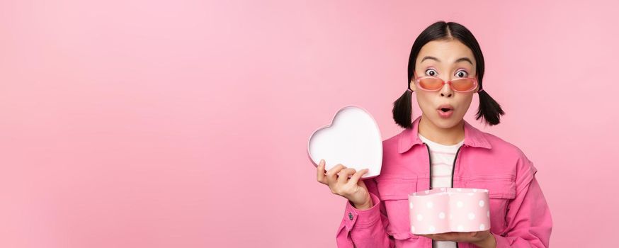 Cute asian girl giving you gift in heart shaped box, kissing and smiling, concept of holiday and celebration, standing over pink background.