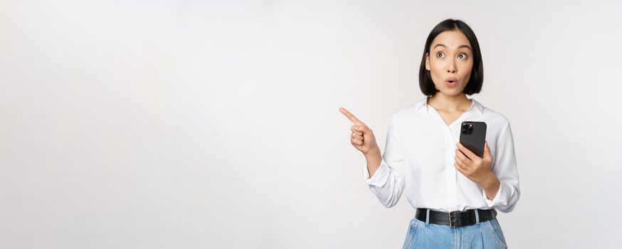 Excited asian girl shopping on mobile app, holding phone and pointing finger left at empty copy space, showing promo text, standing with smartphone against white background.