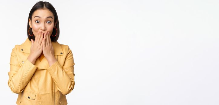 Portrait of excited asian girl looking with interest at camera, amazed reaction at big news or announcement, standing in yellow jacket over white background.