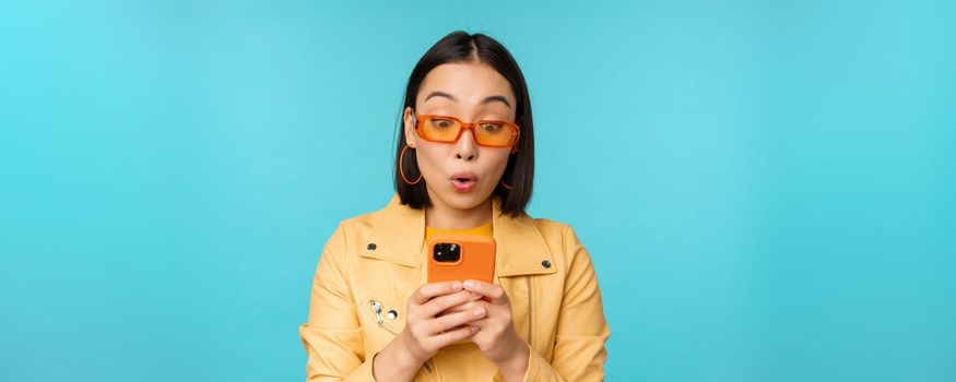 Portrait of asian woman in sunglasses, looking at mobile phone with surprised face expression, standing over blue background. Copy space