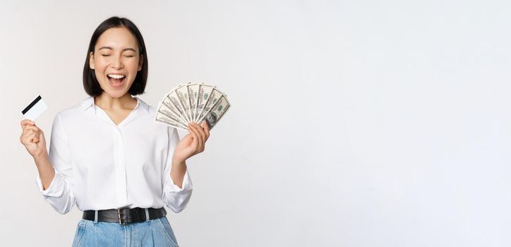 Finance and money concept. Happy young asian woman dancing with cash and credit card, smiling pleased, posing against white studio background.