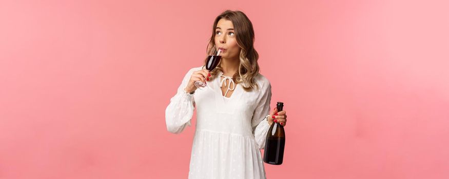 Holidays, spring and party concept. Portrait of carefree independent cute blond woman sipping wine from glass, holding bottle and tasting drink, look up, celebrating with friends, pink background.