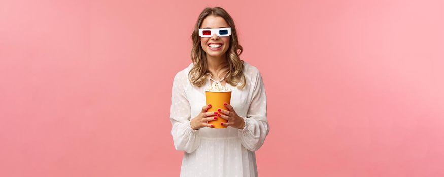 Leisure, going-out and spring concept. Portrait of happy carefree blond girl in white dress enjoying watching movie in 3d, wearing glasses eating popcorn and smiling, attend cinema, pink background.