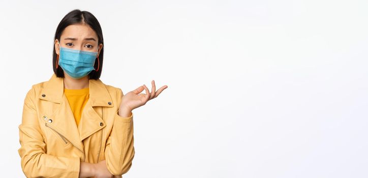 Confused asian woman in medical face mask, looking clueless, puzzled, wearing medical face mask, standing over white studio background.