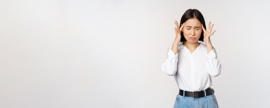 Image of distressed young asian businesswoman, female lady touching head, rubbing temples, has migraine, standing over white background.