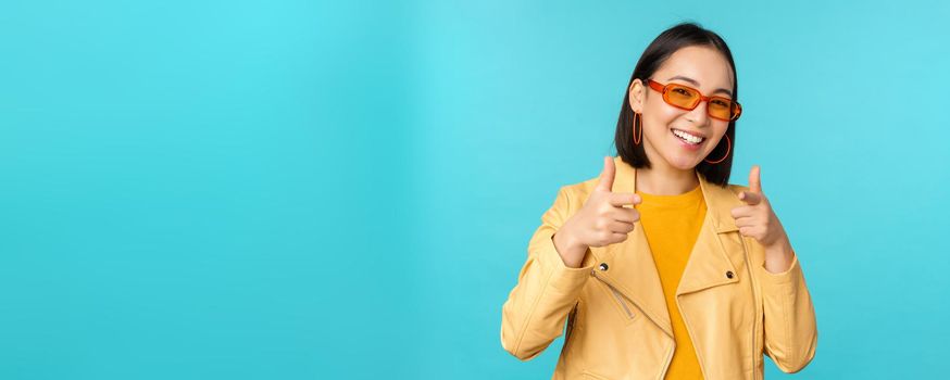 Happy young stylish chinese girl in sunglasses, points fingers at camera with pleased smile, choosing you, congratulating, standing over blue background.