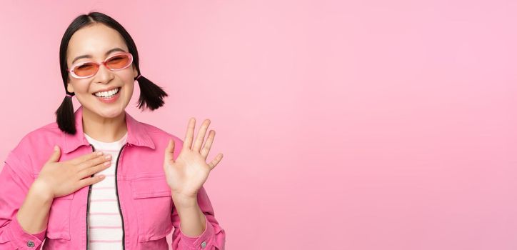 Portrait of friendly asian teen girl in sunglasses saying hello, waves her hand and smiles, greets you, hi gesture, stands over pink background.