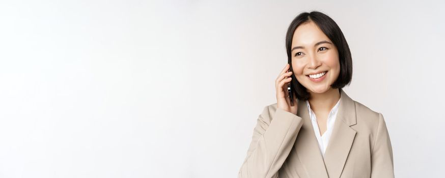 Smiling corporate woman in suit, talking on mobile phone, having a business call on smartphone, standing over white background.