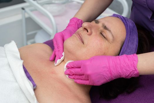A professional beautician cleans the skin on the face with cosmetic pads for a 50-year-old woman