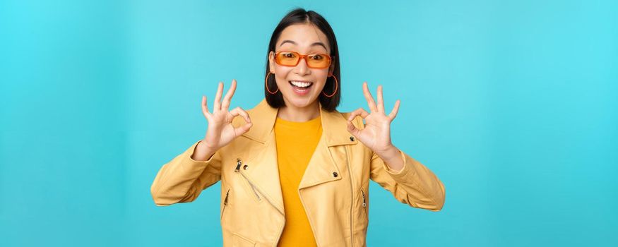 Stylish beautiful asian woman in sunglasses, smiling amazed, showing okay, ok sign, recommending smth, supports excellent choice, standing over blue background.